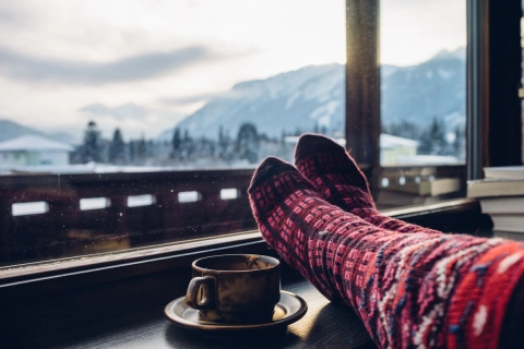 Warme Wintersocken und eine Tasse Tee stehen auf einer Fensterbank und der Blick fällt hinaus auf eine Schneelandschaft.